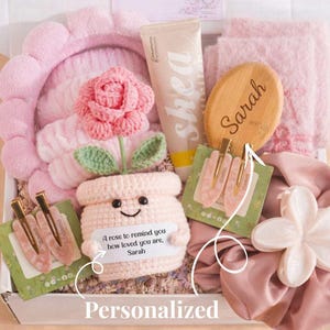 May include: A gift box filled with self-care items in shades of pink. Includes a headband, a tube of shea lotion, a wooden brush with "Sarah" engraved, hair clips, a scrunchie with a flower, and a crochet flower pot with a rose.