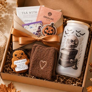 May include: A gift box with a Halloween theme. Inside, a glass jar with a skull design, tea packets, a brown knitted potato with a positive message, and a brown felt coaster with a heart. The box is tied with a gold ribbon.