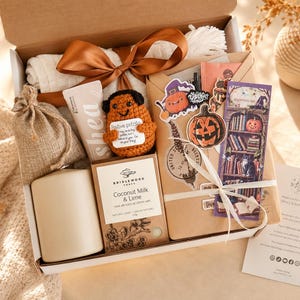 May include: A seasonal gift box with a brown ribbon, containing a candle, a crocheted potato, and a bar of soap. The box also includes stickers and a bookmark with a pumpkin theme, perfect for autumn.