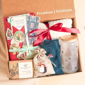 Peut inclure: Un coffret cadeau de Noël avec divers articles. Il comprend une enveloppe rouge avec des autocollants, une serviette blanche nouée avec un ruban rouge, un verre givré avec des motifs de sapins, un article bleu duveteux et un petit sac avec un dessin de pingouin.