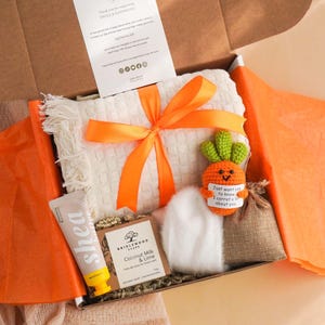 May include: A gift box containing a white fringed blanket tied with an orange ribbon, shea lotion, soap, a small plush carrot, and a burlap bag. The box is lined with orange tissue paper.