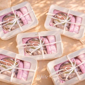 Puede incluir: Cinco neceseres blancos con ventanas transparentes, cada uno atado con una cinta blanca. Las bolsas contienen artículos rosas y beige. La palabra "White" está escrita en la esquina inferior derecha.