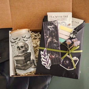 Op de afbeelding: Een geschenkdoos met een kaars met schedelthema, een verpakt pakket en theezakjes. De kaars heeft een schedel- en vleermuisontwerp. Het pakket is zwart met stickers en een groen lint. De theezakjes zijn voorzien van "TEA WITH TAE".