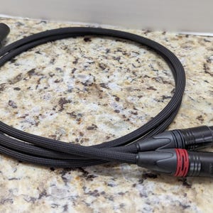 Può includere: Un cavo audio nero con due connettori XLR maschio a ciascuna estremità. I connettori hanno dettagli rossi e neri. Il cavo è arrotolato e poggia su una superficie maculata.