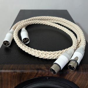 Może przedstawiać: Kremowy kabel audio z białymi złączami, spoczywający na czarnej powierzchni. Kabel ma plecioną teksturę i jest podłączony do głośnika z ciemnym drewnianym wykończeniem i czarnym stożkiem głośnika.