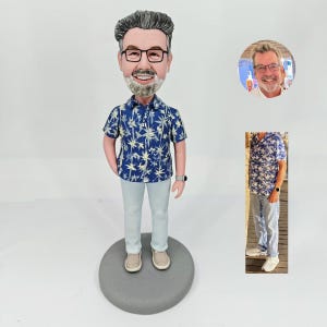 Peut inclure: Figurine personnalisée d'un homme portant une chemise bleue à motifs floraux, un pantalon gris clair et des chaussures beiges. La figurine a des lunettes et une barbe grise. La figurine est sur une base circulaire grise. Deux petites images sont également incluses.