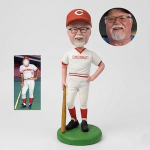 May include: A personalised bobblehead of a man in a Cincinnati Reds baseball uniform, holding a bat. The figure wears a red cap and socks, with the word "Cincinnati" on the jersey. A photo of the man is in the upper right corner.