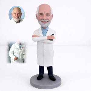 May include: A bobblehead figurine of a smiling, older man with a white beard and hair, wearing a white lab coat, blue shirt, and dark pants. The figurine has its arms crossed and holds eyeglasses. A smaller image of the man is in the upper left.