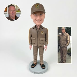 May include: A UPS delivery man bobblehead doll wearing a brown uniform with yellow stripes and a matching cap. The doll has a smiling face and is standing on a gray base. The base reads "UPSMAN 11".