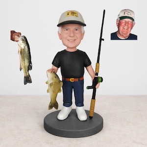 May include: A figurine of a fisherman holding a fish and a fishing rod. The figurine wears a black shirt, blue jeans, and a baseball cap. A second fish is held by a hand in the background. A small photo of a man in a hat is in the background.