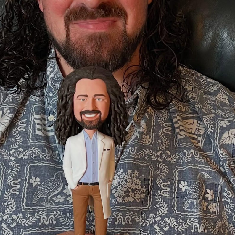 May include: A custom bobblehead figurine of a person with long brown hair and a beard, wearing a white lab coat over a blue shirt and brown pants. The figurine is standing in front of a person wearing a blue and white patterned shirt.