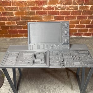May include: A gray 3D printed miniature control console featuring a rectangular monitor with three circular knobs on the right side, positioned above a flat desk surface covered in intricate raised geometric patterns, buttons, and switches.