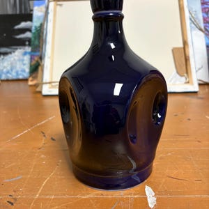 May include: A dark blue ceramic bottle with a glossy finish. The bottle has a unique, rounded shape with two circular indentations on the sides. The bottle's neck is narrow, and it has a small, rounded stopper.