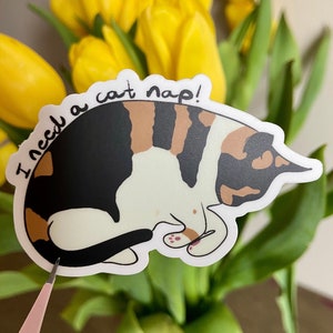 May include: A white sticker with a black outline features a cartoon calico cat curled up sleeping. The text "I need a cat nap!" is written in black cursive around the cat.