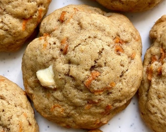 May include: Close-up of six soft, chewy, brown and orange carrot cake cookies with white chocolate chips. The cookies are arranged on a white surface with a speckled pattern.