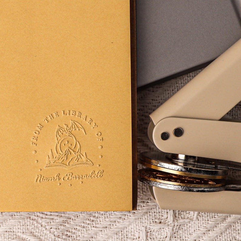 May include: A close-up of a book with a custom embossed stamp. The stamp features a dragon, a crescent moon, and an open book with the text "From the Library of Noah Barradell".