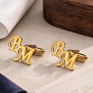 Op de afbeelding: Een paar goudkleurige metalen manchetknopen met de letters B en M in een gestileerd, verbonden scriptlettertype. Elke manchetknop heeft een gepolijst, reflecterend oppervlak en een standaard T-vormig sluitingsmechanisme aan de achterzijde.