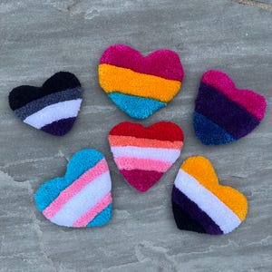 May include: Six heart-shaped crocheted patches in various colors representing different pride flags. The flags include the transgender, non-binary, bisexual, pansexual, asexual, and lesbian pride flags.