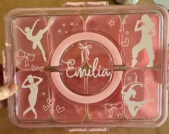 May include: Pink plastic lunch box with multiple compartments. The lunch box has a clear lid and a pink glitter finish. The name "Emilia" is written in white on the front of the lunch box.