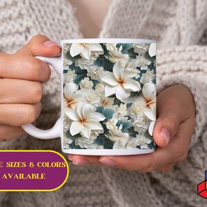 May include: White ceramic mug with a floral design featuring white flowers and green leaves. The mug is being held by a person's hands. The text "MORE SIZES & COLORS AVAILABLE" is visible below the mug.
