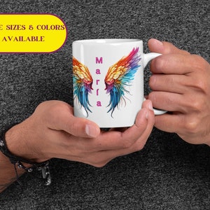 以下が含まれることがあります： カラフルな天使の羽のデザインとピンクの文字で「Marfia」という名前が縦に書かれた白いセラミックマグカップです