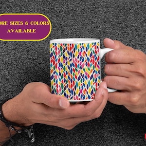 May include: White ceramic mug with a colorful abstract pattern of leaves and swirls. The mug has a white handle. "More Sizes & Colors Available" is written on a purple banner with a yellow border.