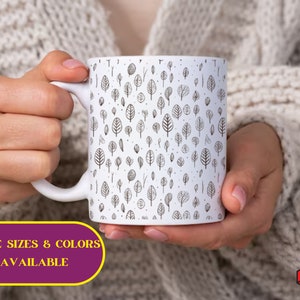 May include: White ceramic mug with a black and white pattern of small trees. The mug is being held by a person's hands. The text "MORE SIZES & COLORS AVAILABLE" is printed below the mug.
