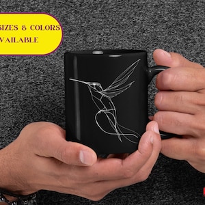 May include: Black ceramic mug with a white line drawing of a hummingbird. The mug has a handle on the right side. The text "MORE SIZES & COLORS AVAILABLE" is in a yellow rectangle at the top of the image.