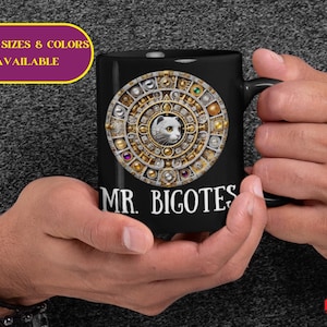 May include: Black ceramic mug featuring a detailed, circular design with a cat's face at the center, surrounded by gold and jewel-toned accents. The text "MR. BIGOTES" is printed below the design. The mug is held in two hands.