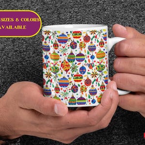 May include: A white ceramic mug featuring a colorful pattern of Christmas ornaments in various shapes and sizes. The mug is held by hands, and the text "MORE SIZES & COLORS AVAILABLE" is displayed on a yellow oval.