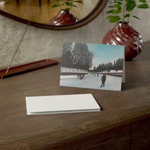 May include: A folded greeting card with a winter scene painting, featuring people ice skating on a frozen waterway. The card is displayed on a wooden surface, with a blank card in front. A decorative vase and mirror are in the background.