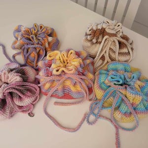 Pode incluir: Cinco sacos de cordão de crochê em várias cores, incluindo rosa, roxo, amarelo, azul e marrom. Os sacos são feitos com um padrão listrado e têm um topo franzido.