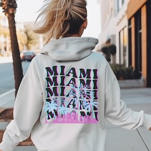 Puede incluir: Una sudadera con capucha blanca con un estampado gráfico negro de la palabra "Miami" repetida cuatro veces con palmeras y un horizonte de ciudad en el centro. El gráfico tiene un esquema de color azul y rosa.