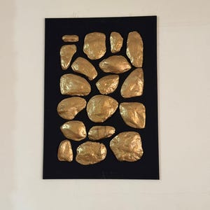 Può includere: Un'opera d'arte murale rettangolare nera con una collezione di pietre dorate di forma irregolare. Le pietre sono disposte in un motivo apparentemente casuale, creando un effetto strutturato e tridimensionale. L'opera è montata su una parete bianca.