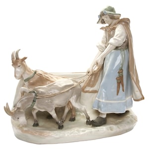 Puede incluir: Figura de porcelana que representa a una mujer con un vestido azul y una capa, guiando a dos cabras enganchadas a un carro. Las cabras son de color gris claro y marrón, y la mujer lleva un sombrero. La figura está sobre una base blanca.