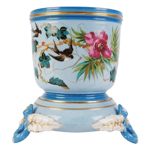 Puede incluir: Un macetero de porcelana azul y blanco con ribete dorado y diseño floral y de pájaros. El macetero tiene tres patas con forma de hoja.