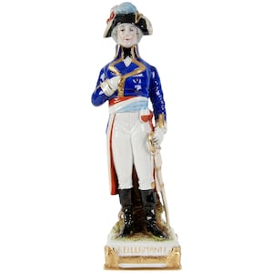 Op de afbeelding: Een porseleinen beeldje van een militair figuur in een blauwe jas met gouden biezen, een witte broek en zwarte laarzen. Het figuur houdt een zwaard vast en staat op een voetstuk met de naam "KELLERMANN" in gouden letters. De hoed heeft een veer.