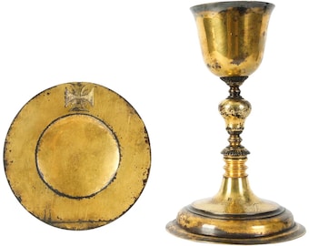 May include: A gold-colored chalice and paten set. The chalice has a detailed stem and a flared base. The paten is a round plate with a cross design in the center. Both items show signs of wear.