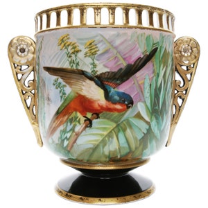 Könnte beinhalten: Eine Porzellanvase mit goldenen Akzenten und einer bemalten Szene eines Papageis in tropischer Umgebung. Die Vase hat zwei Henkel mit Blumendesigns und einen schwarzen Sockel.