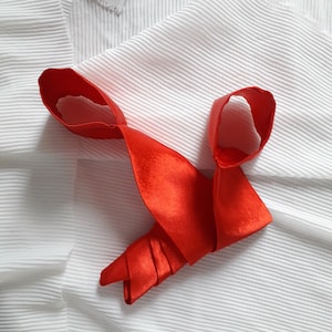 May include: A red satin tie folded and arranged on a white fabric background.