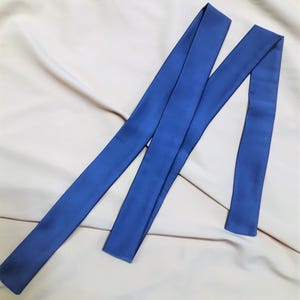 Puede incluir: Dos largas y estrechas fajas de tela azul cobalto están dispuestas diagonalmente sobre un fondo neutro blanquecino. Las fajas parecen estar hechas de un material liso, similar al satén, con un ligero brillo. La tela está doblada y drapeada.