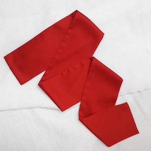 Puede incluir: Una faja roja vibrante, probablemente de satén, se muestra sobre un fondo blanco texturizado. La faja está doblada y drapeada, mostrando su longitud y textura suave. El color brillante la hace adecuada para varios eventos.