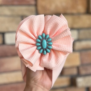 May include: A peach-colored baby headband with a textured fabric design. The headband features a turquoise and silver-colored decorative centerpiece. The accessory is held up against a brick wall background.