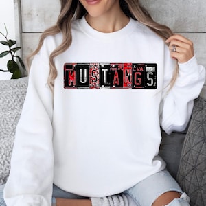 Puede incluir: Sudadera blanca con un gráfico rojo y negro de la palabra "MUSTANGS" formado por placas de matrícula.