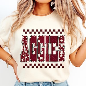 Op de afbeelding: Crèmekleurige sweatshirt met het woord "AGGIES" in bordeauxrood en wit. De letters hebben ster- en patroonontwerpen. De boven- en onderkant van het ontwerp hebben een bordeauxrood en wit geblokt patroon.