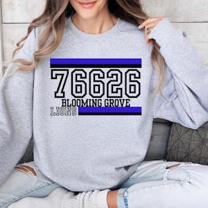 Puede incluir: Una sudadera gris claro con el texto "76626 BLOOMING GROVE LIONS" en negro y azul. La sudadera tiene mangas largas y cuello redondo. El texto está en un estilo de letras de bloque.