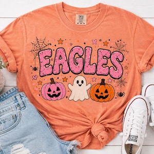Peut inclure: Un t-shirt orange à manches courtes avec le texte EAGLES en grandes lettres roses. Le motif comprend une citrouille rose, un fantôme blanc, une citrouille orange, des toiles d'araignée, des araignées et des petites étoiles sur le tissu.