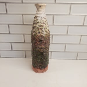 May include: A ceramic vase with a textured, tree bark-like design in shades of brown, green, and white. The vase has a narrow neck and a rounded base.