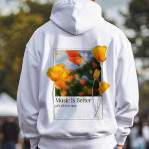 May include: White hoodie featuring a floral design with yellow and orange flowers. The graphic includes the text "Music Is Better" and "RÜFÜS DU SOL" in a rectangular frame. The hoodie has a hood and long sleeves.