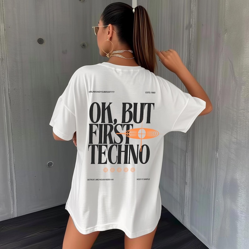 May include: White t-shirt with the text "OK, BUT FIRST TECHNO" in black block letters. The shirt has an orange and white graphic design. The text "DETROIT, MICHIGAN 48201-44" is printed at the bottom of the shirt.